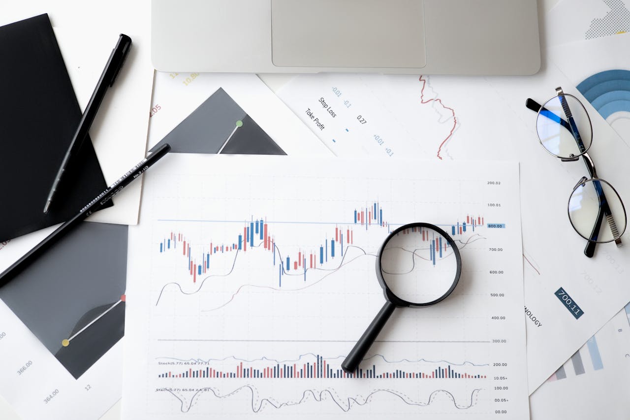 hero-img-01 Flat lay of stock market analysis documents with magnifying glass, pens, and glasses.